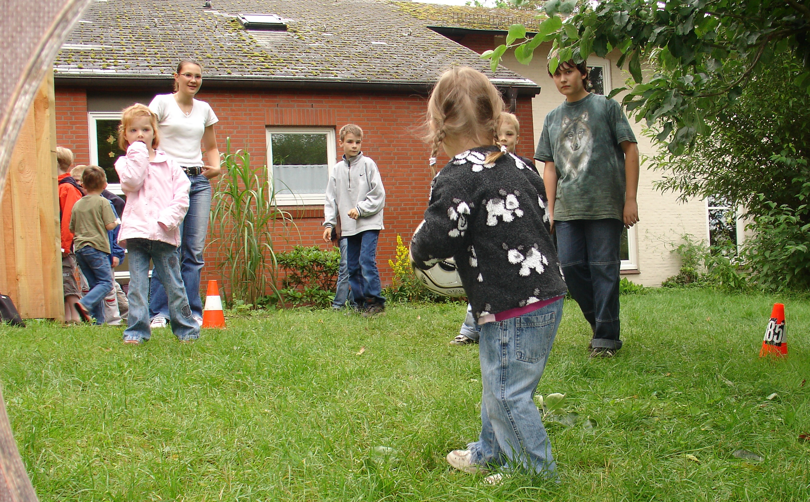 Kinder und Erwachsene im Freien auf einer Wiese vor einem kleinen Haus.