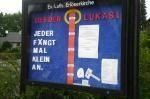 Schwarze Schautafel mit blauem Hintergrund, Text und Aushängen vor einer Kirche.