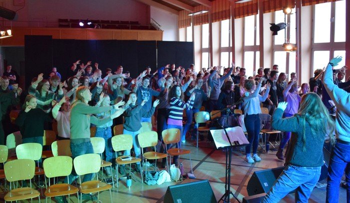 Große Gruppe singender oder tanzender Menschen in einem beleuchteten Raum.