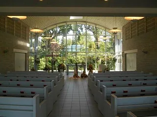 Heller Innenraum einer Kapelle mit Holzbänken und einem Altar vor einer großen Glasfront.